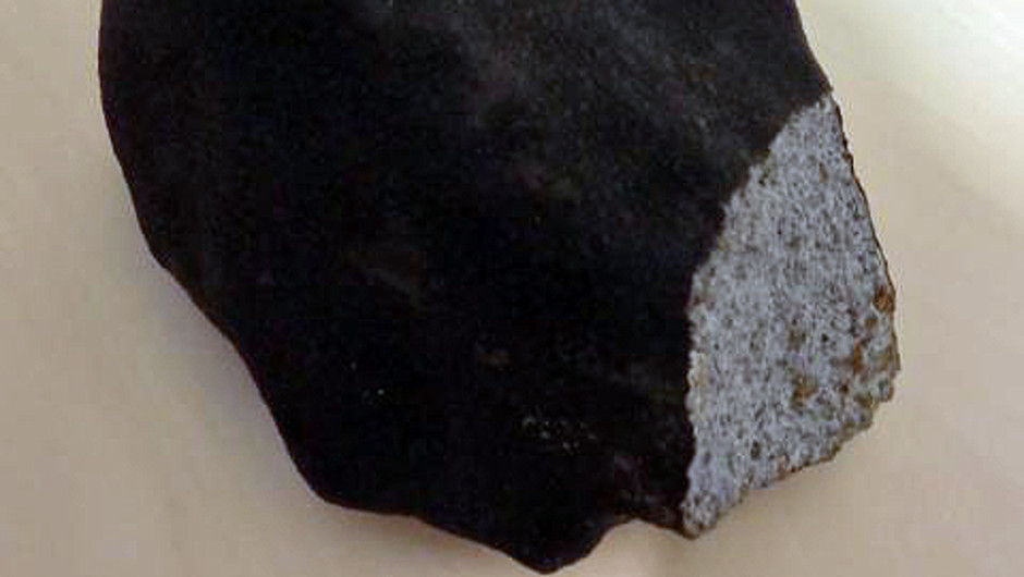 Ett stycke av stenmeteoriten Hessle från Naturhistoriska riksmuseets samling. Nere till höger syns en färsk avslagen yta, i övrigt den brunsvarta smältskorpan. 