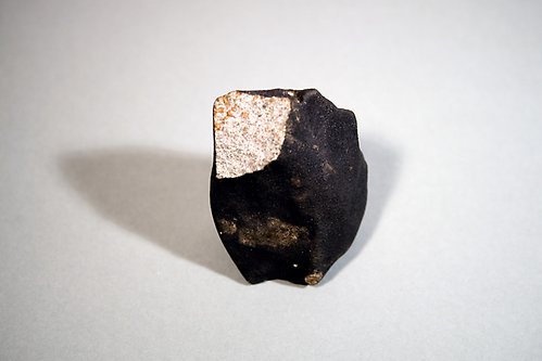 En meteorit med klassiskt svärtad utsida. Ett hörn av stenen saknas och där syns ett ljusare beige innanmäte.