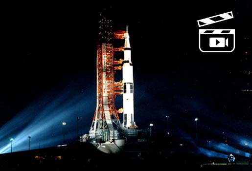 Apollo 14 på startplattan. Det är mörkt, raketen är belyst med strålkastare. I övre högra bildhörnet är det en vit ikon som föreställer en filmklappa.