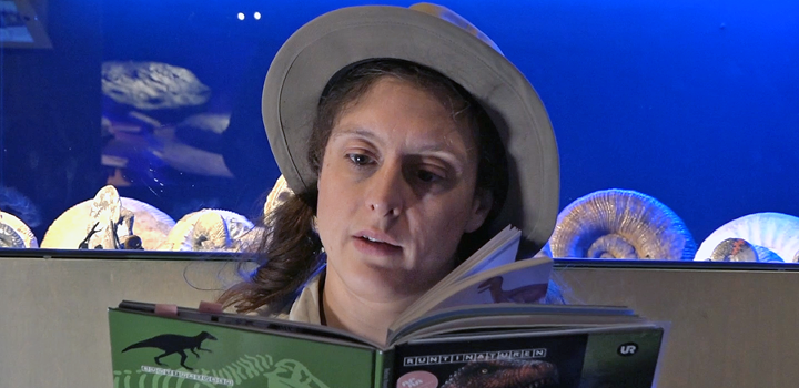 Dino-Doris i hatt sitter framför en monter med ammoniter och läser i en bok.
