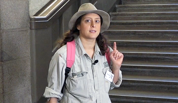 Dino-Doris i hatt, khakifärgade kläder med rosa ryggsäck står i museets trappa med ett lyft pekfinger.