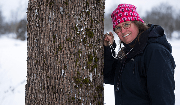 En kvinna i rosa mössa håller en lupp mot en trädstam mot en bakgrund av ett vinterlandskap.