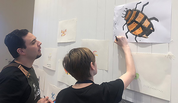 En man tittar på när ett barn ritar på en bild av en skalbagge som sitter på en vägg med andra teckningar.