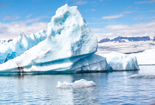 I förgrunden hav och ett isberg, i bakgrunden syns snötäckt mark och ett blånande långsträckt bergsmassiv.