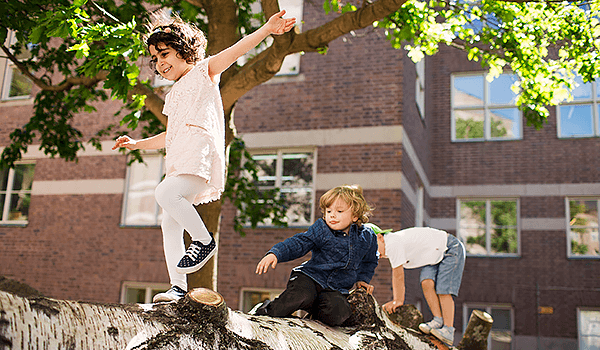 Three children climb on a tree trunk with a brick building in the background.